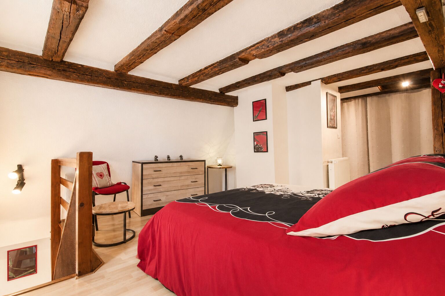A bedroom with a wooden ceiling, a bed with a red and black bedspread, a wooden dresser, and a chair with a red cushion.