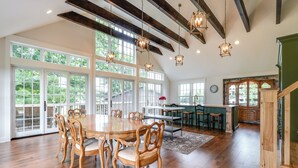 A spacious dining area with a large wooden table and chairs, surrounded by large windows offering a view of greenery.