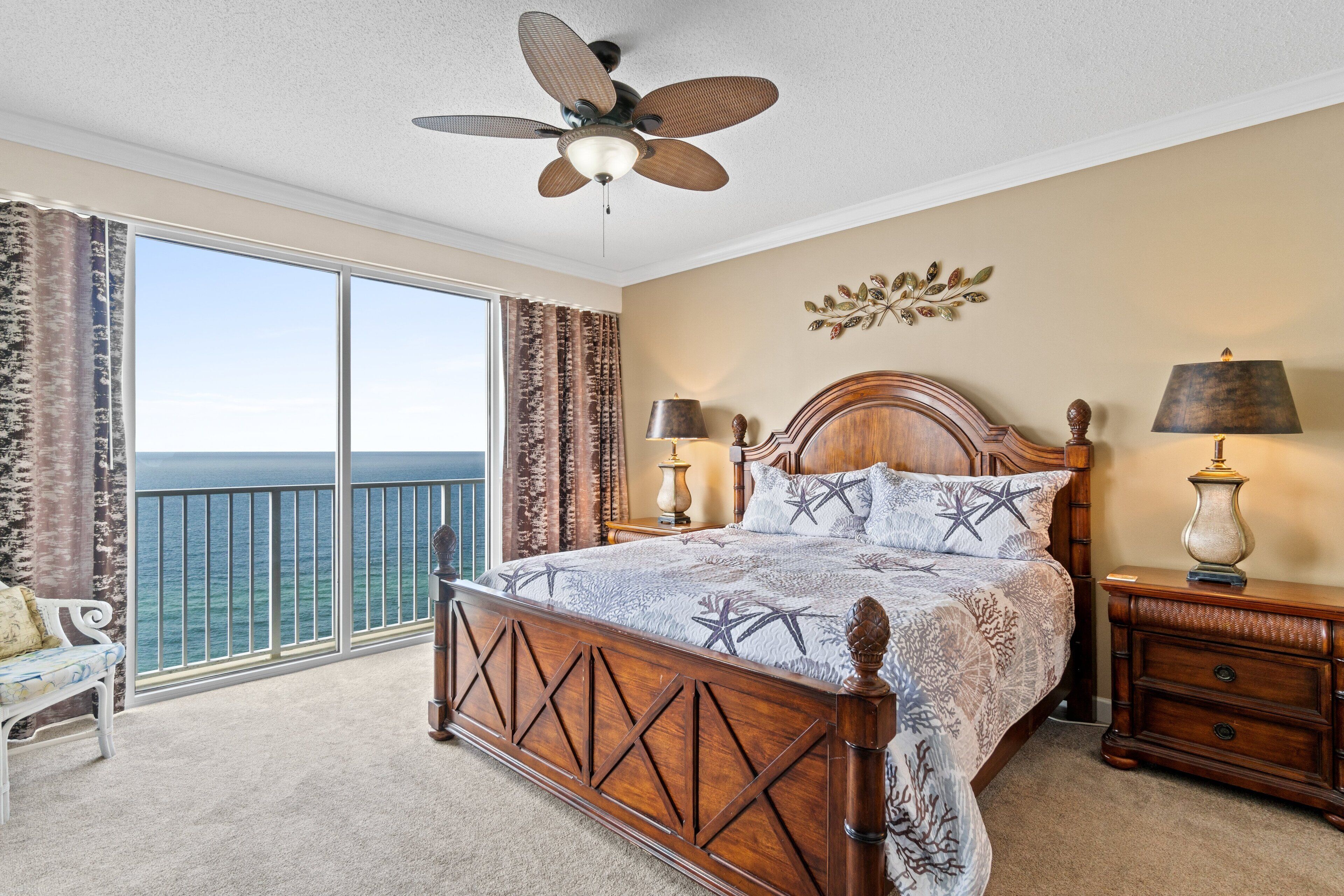 A bedroom with a large bed, a ceiling fan, a nightstand, and a view of the ocean.