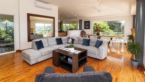 A spacious living room with a large sectional sofa, a coffee table, and a dining area with a view of greenery. - Casa Diamante - Top Rated Vacation Home in Manuel Antonio! (Quepos)