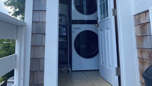 Une buanderie équipée de deux machines à laver à chargement frontal superposées, d’un sèche-linge et d’une porte donnant sur un porche abrité.