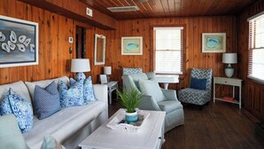 Une pièce chaleureuse aux murs en bois, avec un canapé blanc orné de coussins à motifs bleus, un fauteuil gris et une petite table blanche sur laquelle est posée une plante.