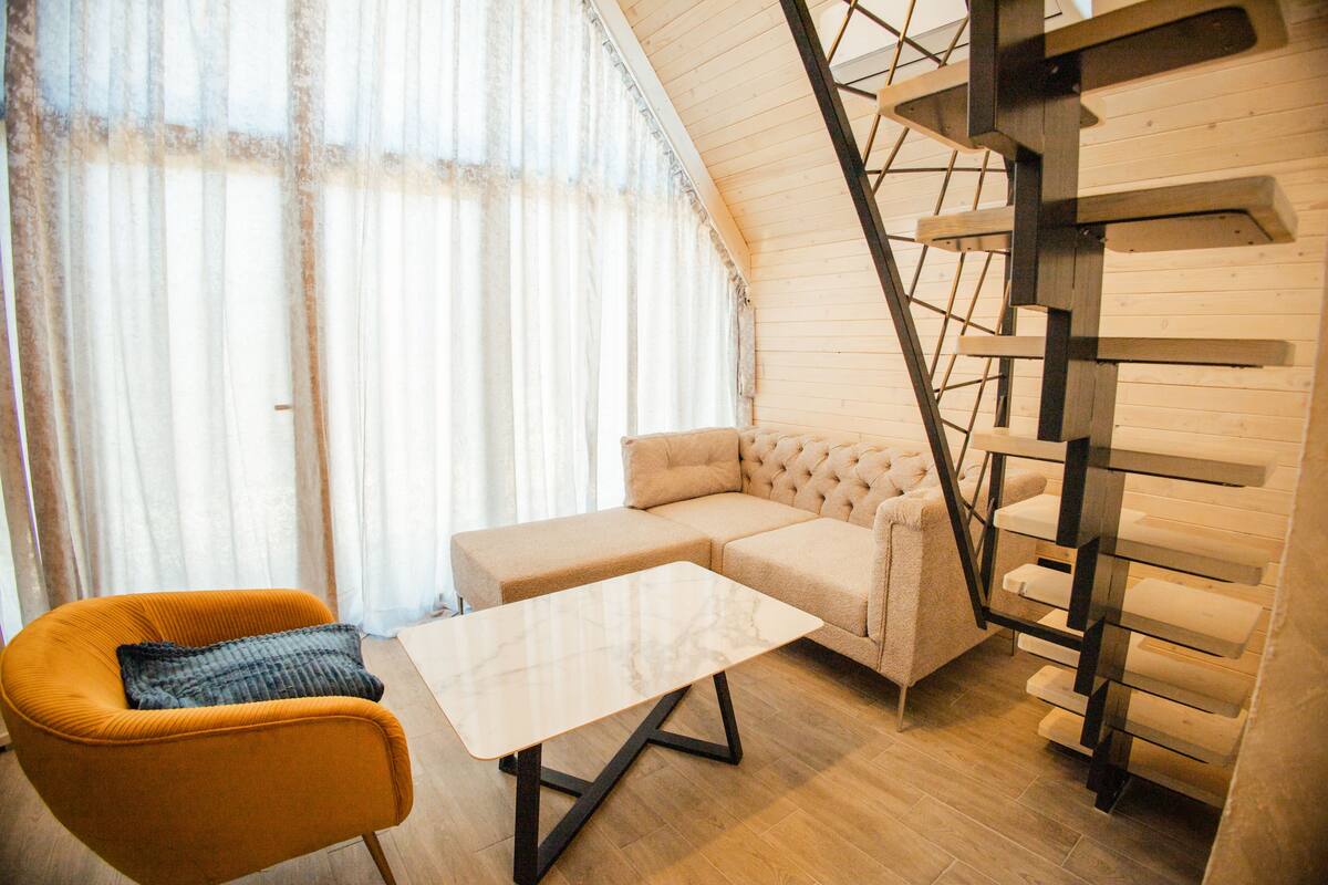 A modern living room with a sofa, a small table, and a chair. There is a staircase with wooden steps and metal railings.