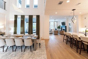 Ein moderner Essbereich mit einem langen Tisch, gedeckt für sechs Personen, eine Küchenzeile mit Barhocker und ein Wohnbereich mit Sofa und Fernseher.