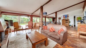 Een ruime woonkamer met een houten salontafel, een bruine bank met kussens, en een televisie op een houten dressoir. Grote ramen zorgen voor natuurlijk licht en uitzicht op groen buiten.