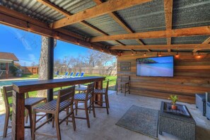 A covered outdoor area with a wooden table and chairs, a TV screen, and a view of a swimming pool and backyard. - Crystal Cow Haus Luxury Home-Pool-HotTub-Near Main (Fredericksburg)