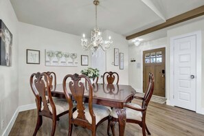 Une salle à manger avec une table et des chaises en bois, un lustre, des œuvres d’art encadrées accrochées aux murs et une porte en bois à l’arrière-plan.