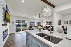 A modern kitchen with a large island, a fireplace, and a dining area with a sofa and chandelier. - Crystal Cow Haus Luxury Home-Pool-HotTub-Near Main (Fredericksburg)