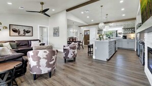 A spacious living room with a dark brown leather sofa, a white coffee table, and a patterned armchair. A ceiling fan and pendant lights are visible.