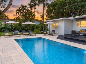 A swimming pool with lounge chairs and umbrellas, surrounded by greenery and a building with a white facade.