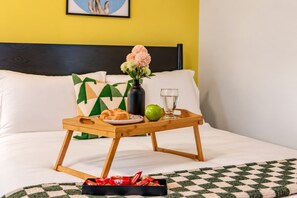 Ein Bett mit Tablett, das ein Croissant, einen Apfel und ein Glas Wasser trägt, dazu ein Blumenstrauß und ein Kissen mit geometrischem Muster im Hintergrund.
