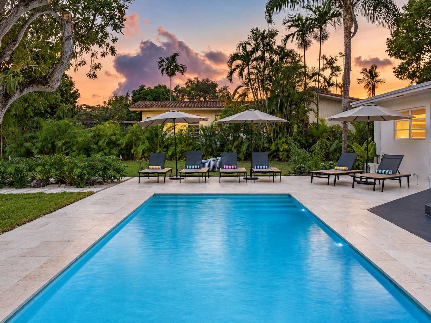 A rectangular swimming pool with lounge chairs and umbrellas, surrounded by lush greenery and palm trees, with a house in the background.