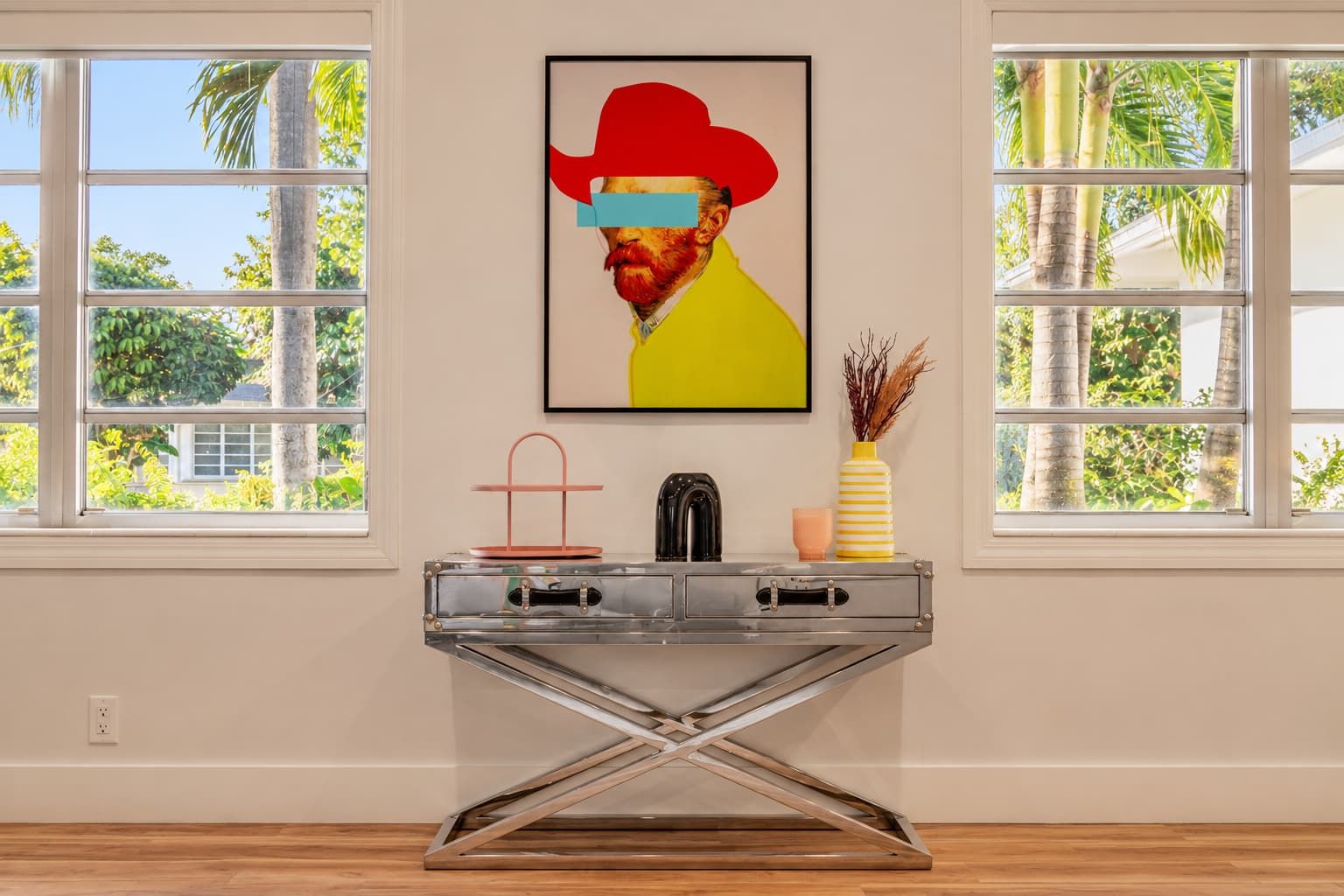 Een modern interieur met een metalen console tafel, een ingelijste afbeelding en uitzicht op palmbomen door grote ramen.