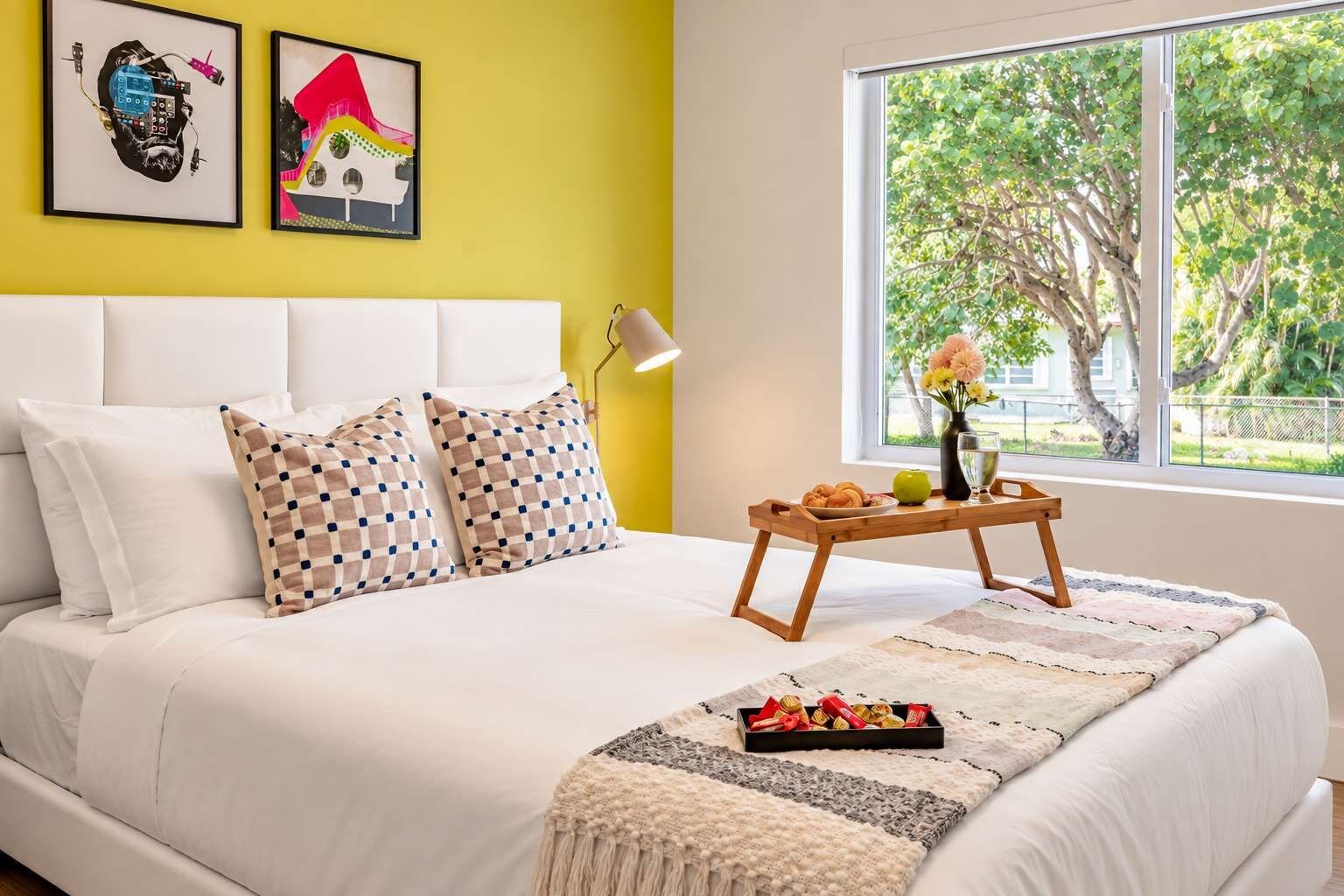 Een bed met witte lakens, geruit kussens en een dienblad met snacks.