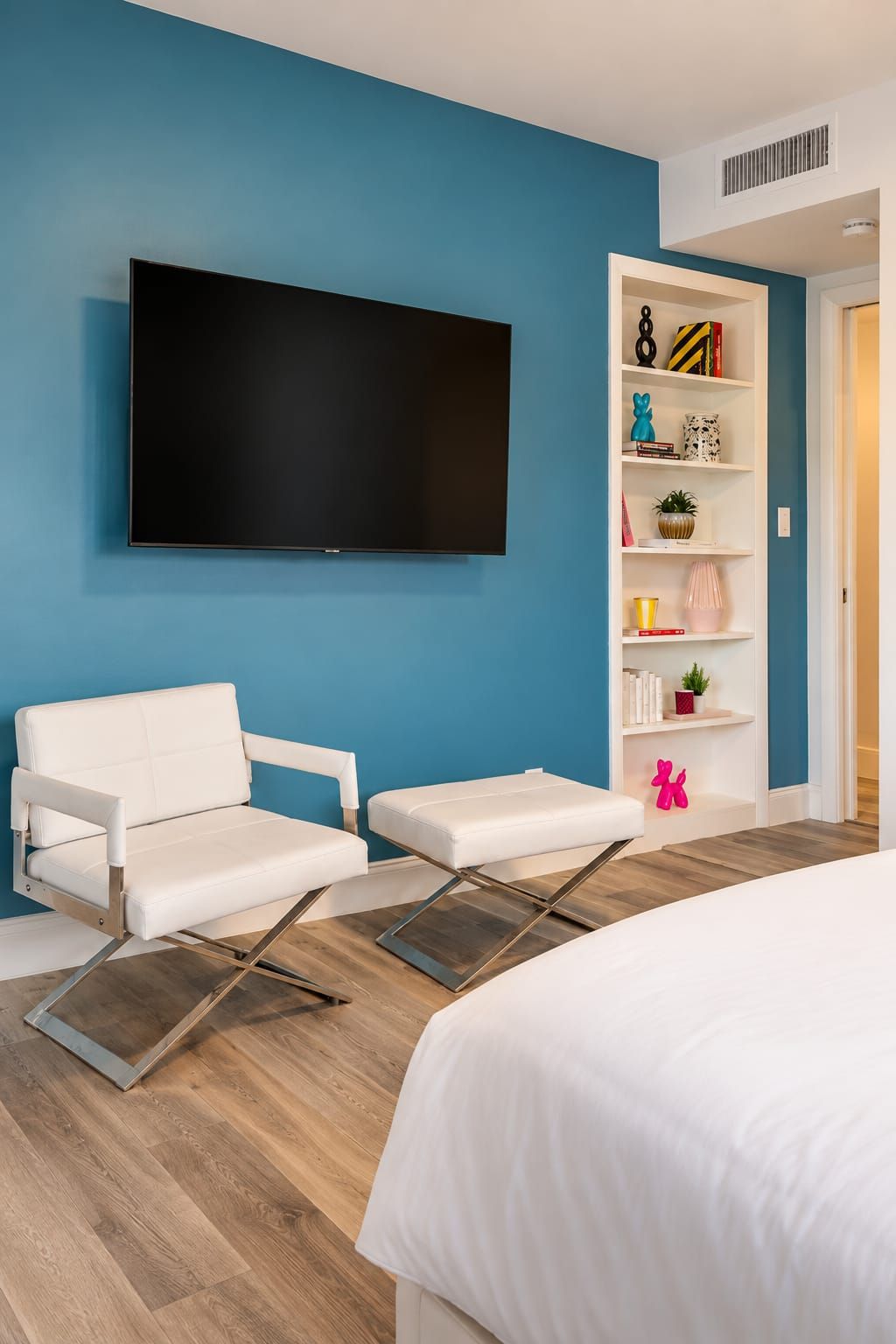 Ein Hotelzimmer mit einem Bett, einem weißen Sofa, einem kleinen weißen Tisch, einem an der Wand befestigten Flachbildfernseher und einem Bücherregal mit diversen Gegenständen.