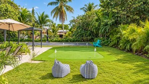 Une pelouse bien entretenue avec un green d’entraînement au golf, deux sacs de jeu à rayures, une chaise bleue et un kiosque.