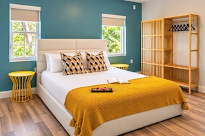A bedroom with a bed, a yellow throw blanket, a bedside table, a wooden wardrobe, and a window with a view of greenery.