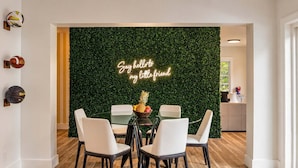 Un espace repas avec une table ronde en verre, des chaises blanches, un panneau lumineux au néon et un mur vert.