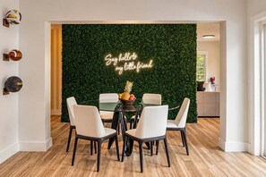 A dining area with a round glass table, white chairs, a neon sign, and a green wall.