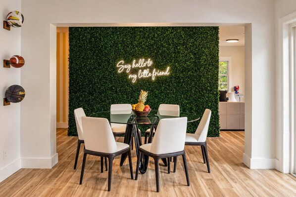 A dining area with a round glass table, white chairs, a neon sign, and a green wall.