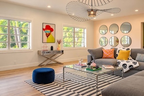 A modern living room with a sofa, coffee table, and a blue ottoman. There are windows with a view of greenery outside.