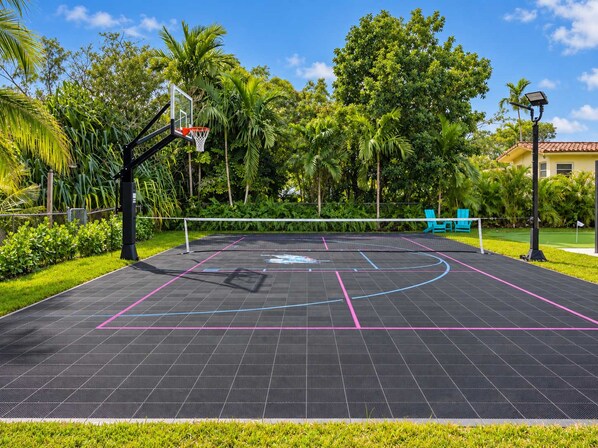 An outdoor basketball court with a net, surrounded by lush greenery and a clear sky.