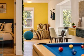 A room with a pool table, a bed with patterned pillows, a dining area with chairs, and a window with greenery outside.