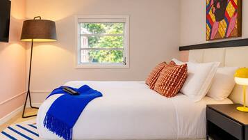 A hotel room with a bed, a nightstand, a lamp, a window with a view of greenery, and a painting on the wall.