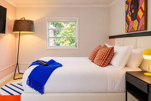 Quarto de hotel com cama, mesa de cabeceira, abajur, janela com vista para o verde e um quadro na parede.