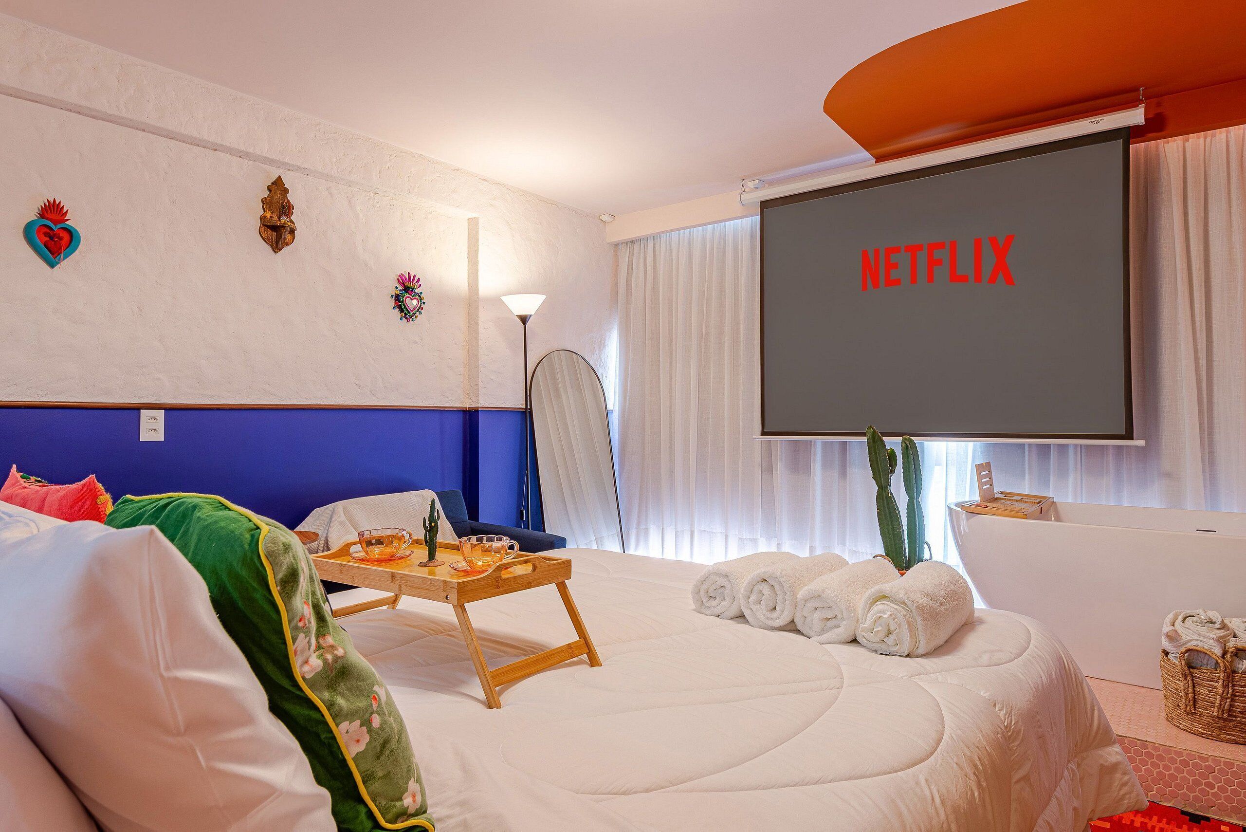 A hotel room with a bed, a desk with a tray, a cactus, and a large screen displaying 'NETFLIX'.