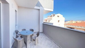 Un balcon avec une table ronde et des chaises en osier, donnant sur un quartier résidentiel avec des immeubles et un ciel bleu dégagé.