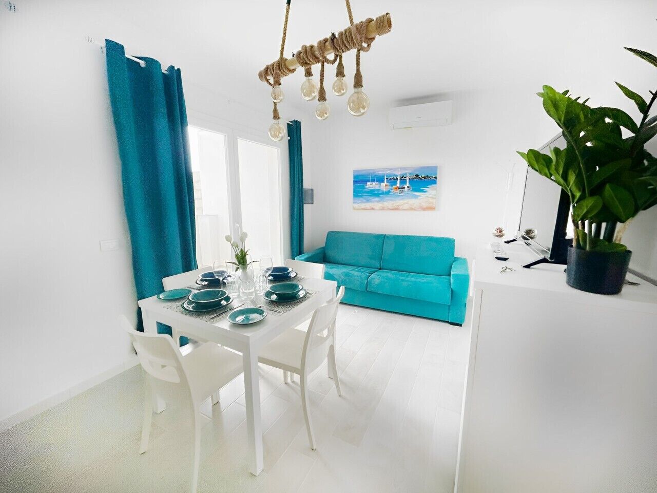 Ein helles, modernes Wohnzimmer mit einem türkisfarbenen Sofa, einem Esstisch gedeckt mit Tellern und Gläsern sowie einer Hängelampe.