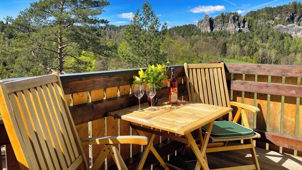 A wooden deck with a table and chairs, overlooking a forested landscape with rocky formations.