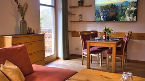 A living room with a wooden dining table, purple upholstered chairs, a wooden sofa, a wooden cabinet, a large window, and a wall-mounted TV.