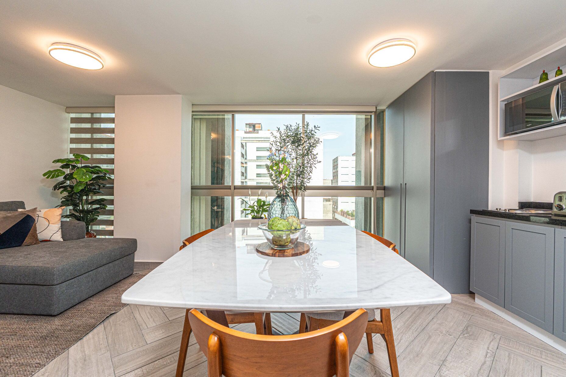 A modern kitchen with a marble countertop, a dining table with chairs, and a view of buildings through large windows.