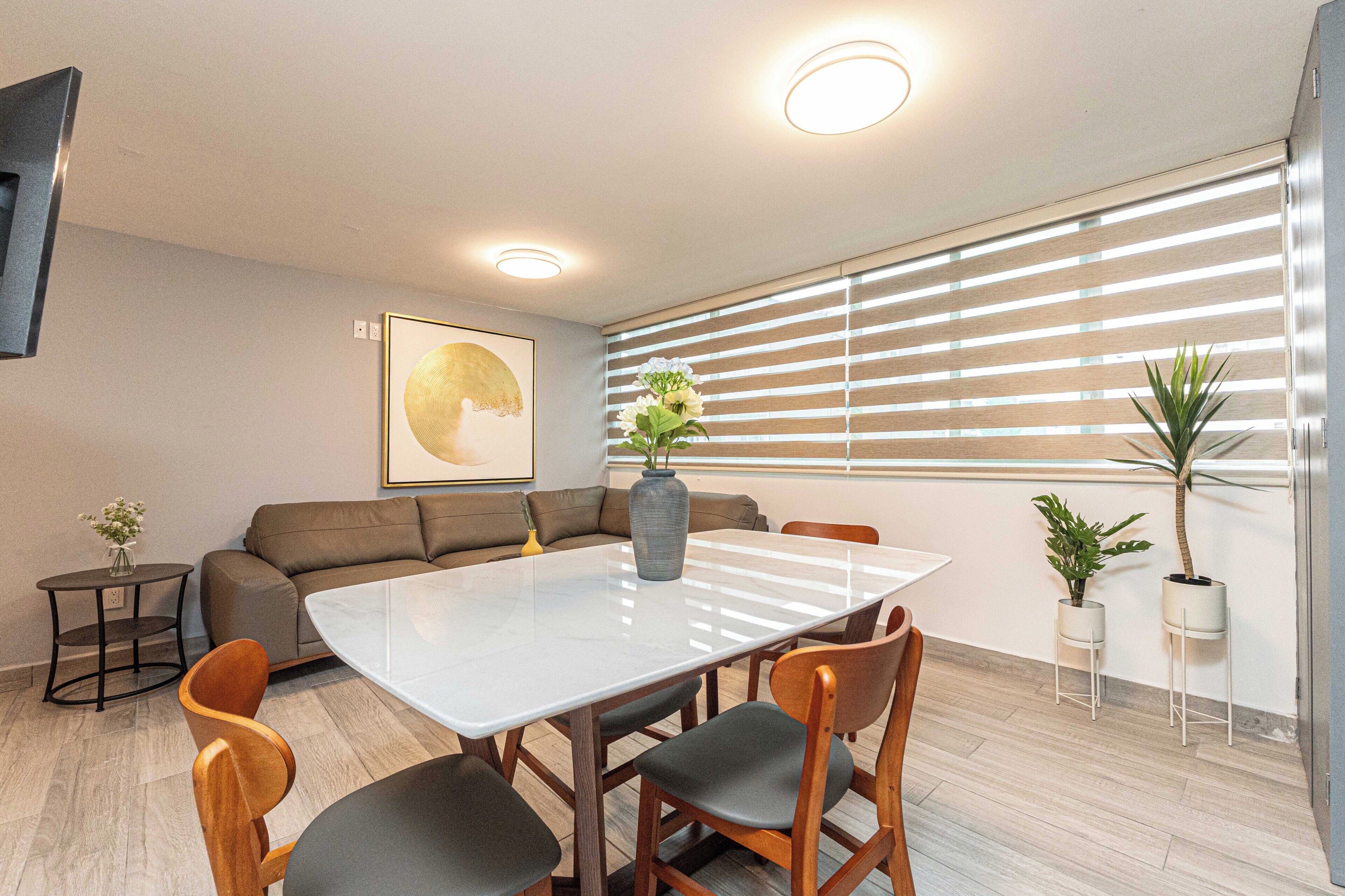 A modern living room with a sofa, a dining table with chairs, and a large window with blinds.