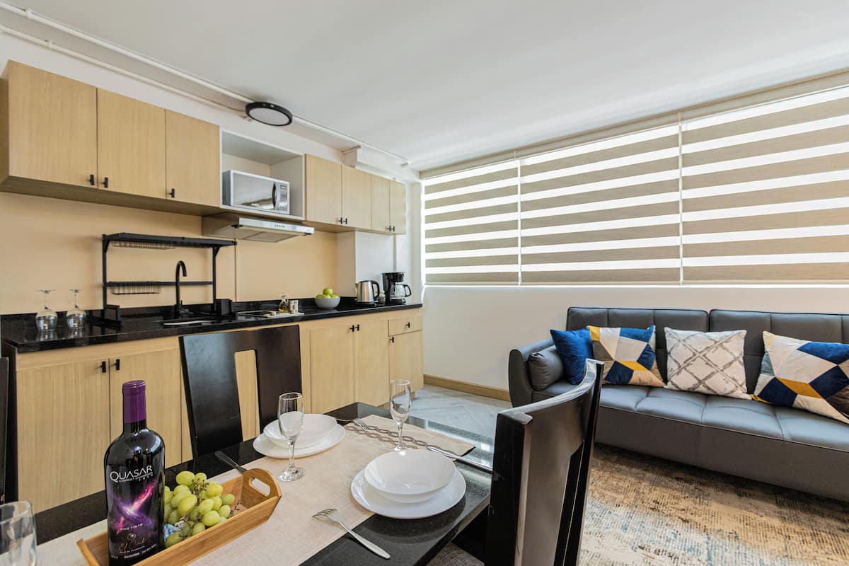 A modern living room with a grey sofa, a dining table set with plates, glasses, and a bottle of wine. There is a kitchen area with wooden cabinets and a microwave.