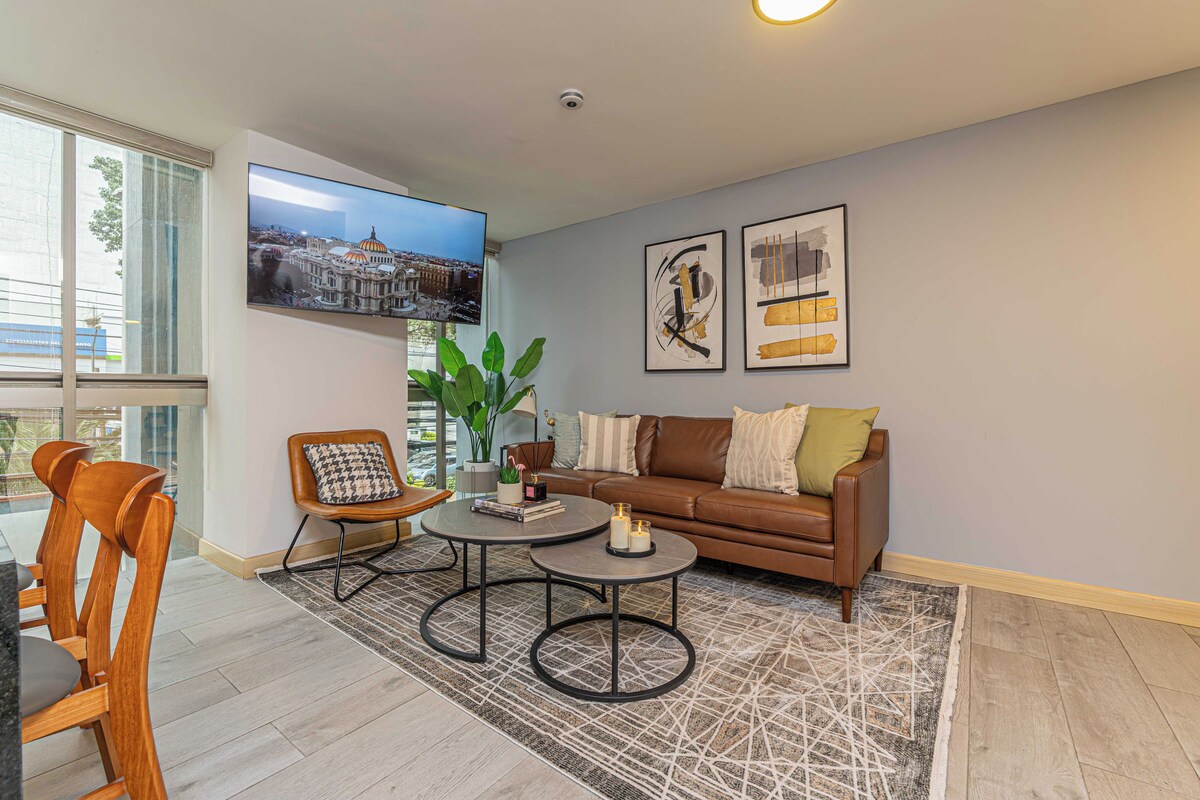 A modern living room with a sofa, coffee table, and chairs, a flat-screen TV mounted on the wall, and artwork on the wall.