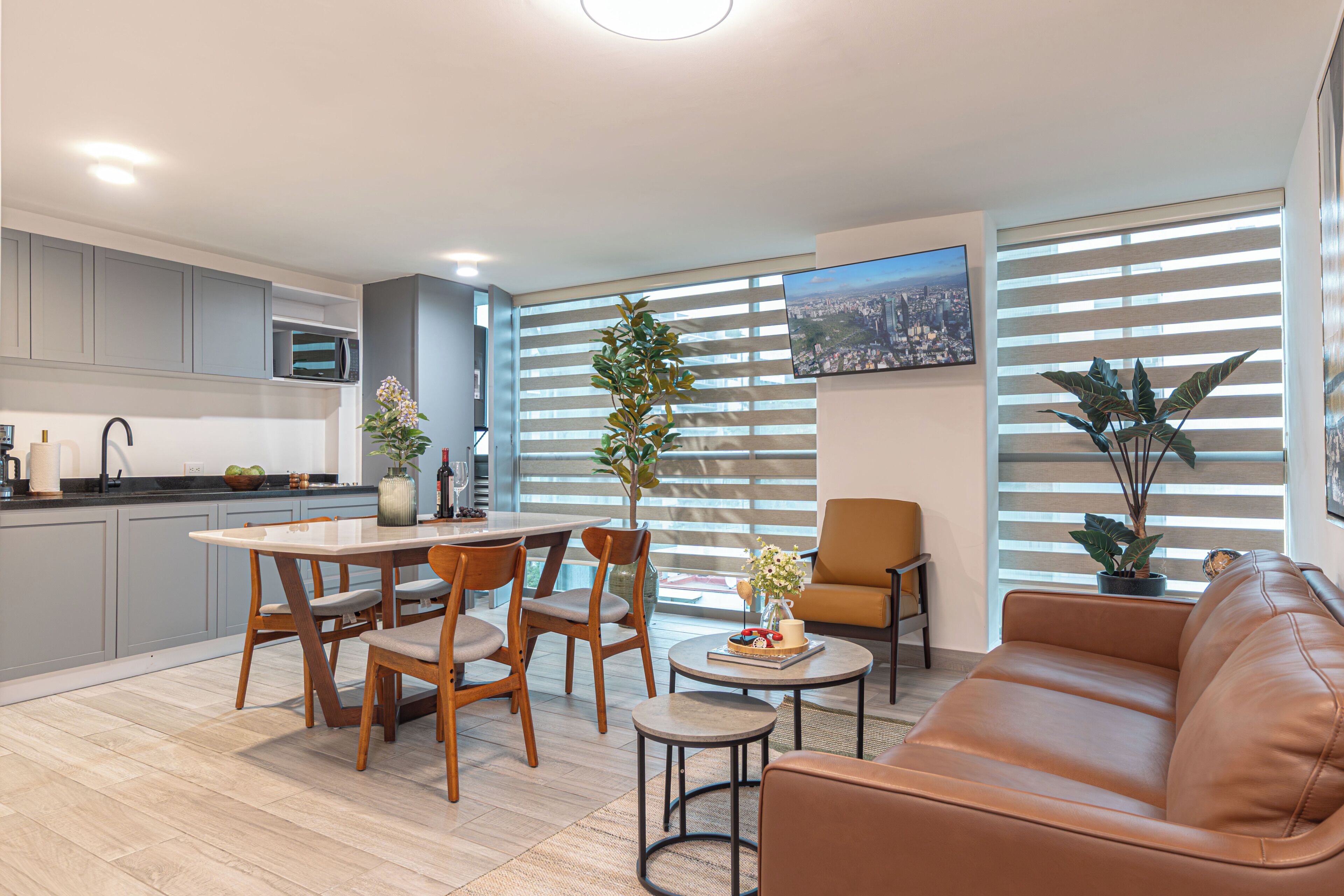 Una sala de estar moderna con un sofá de cuero marrón, una mesa de comedor con sillas, una zona de cocina con armarios grises y un ventanal con persianas.
