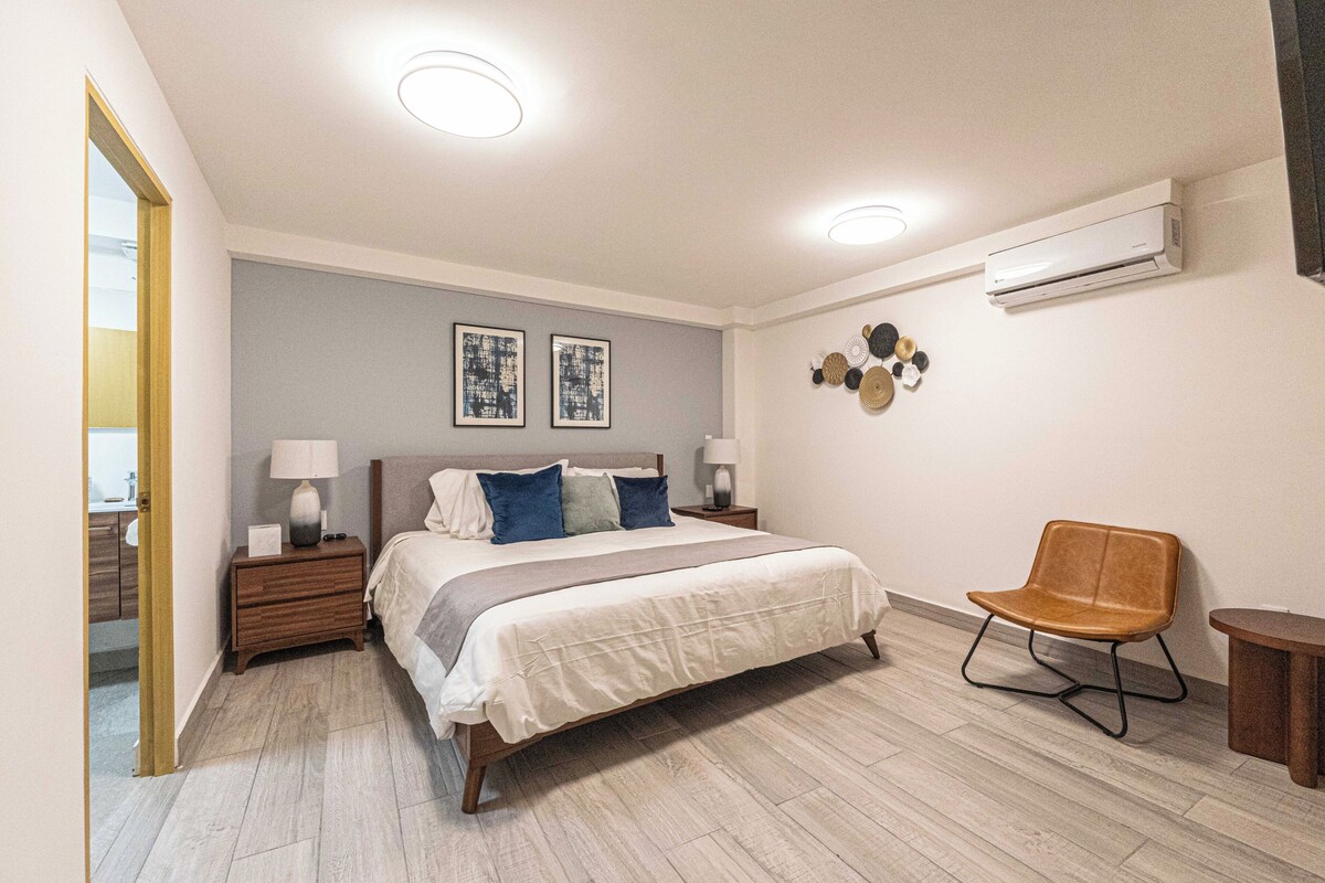 A bedroom with a bed, bedside tables, a chair, a wall-mounted air conditioner, and two framed pictures.