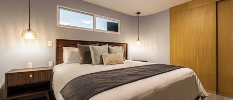 A modern bedroom with a large bed, two pendant lights, a nightstand, a wooden wardrobe, and a window above the bed.