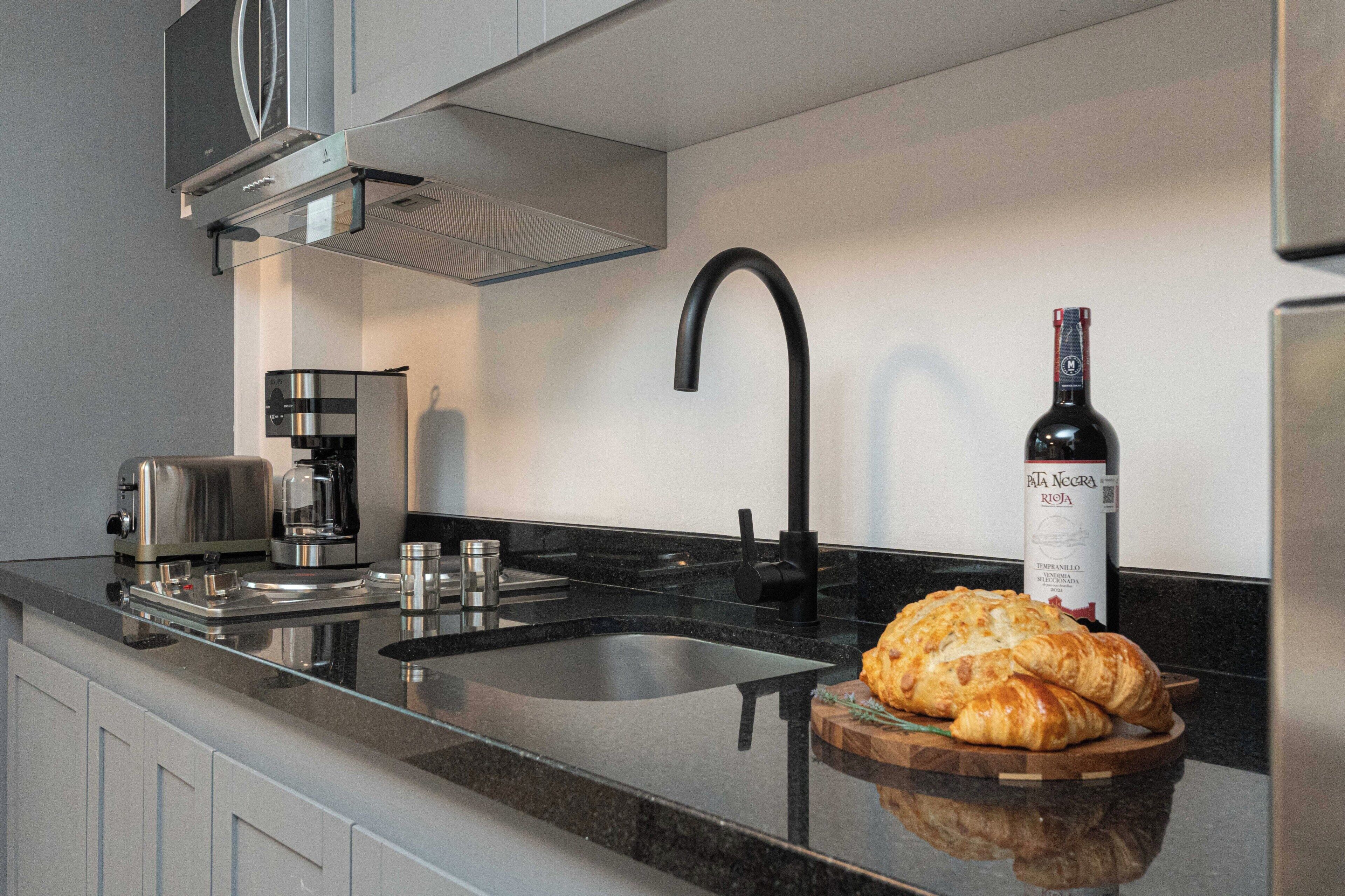 A modern kitchen with a stainless steel range hood, a black faucet, a wine bottle, and a wooden cutting board with croissants.