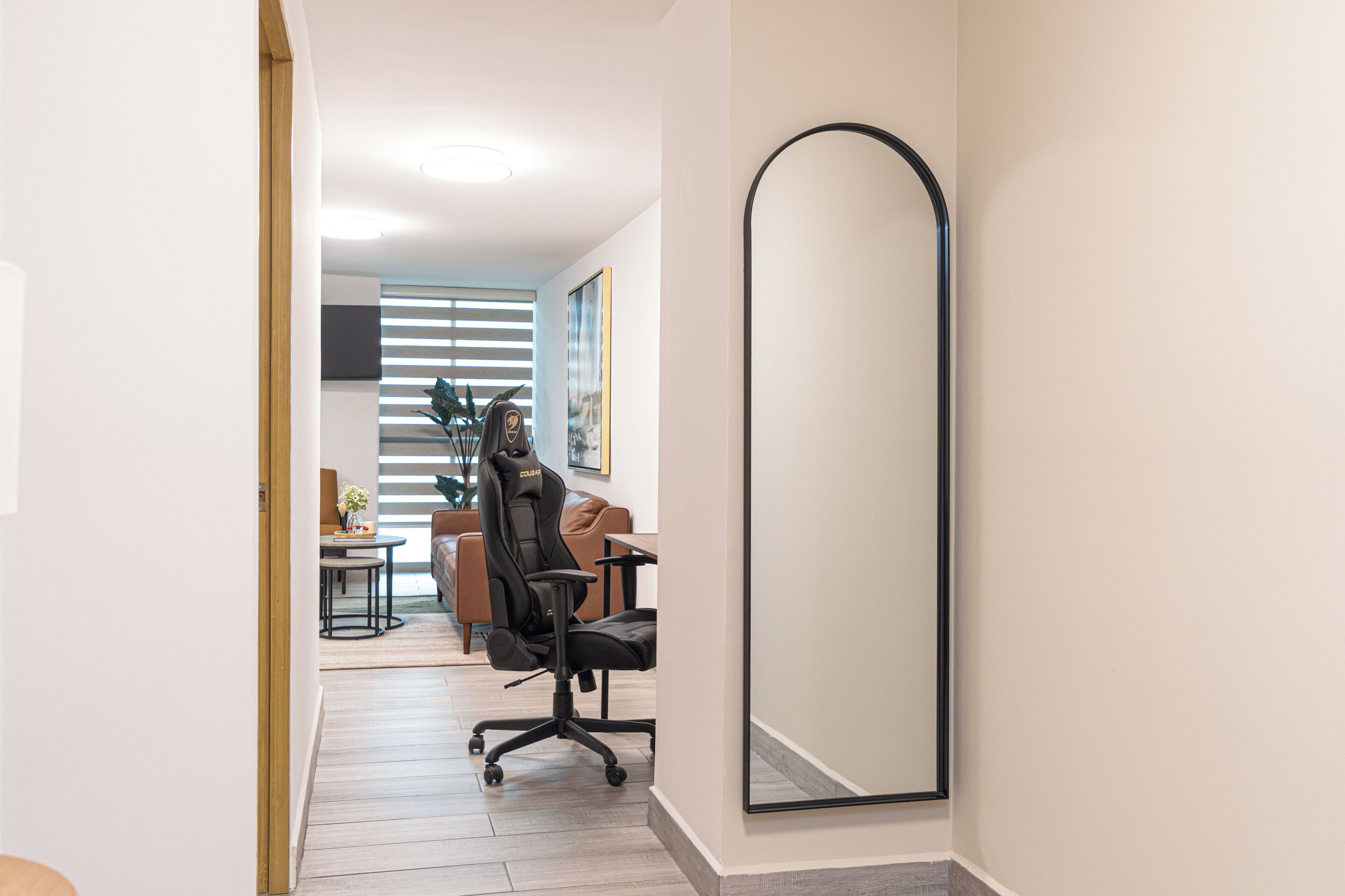 A hallway with a large mirror, a black office chair, a brown sofa, and a dining table with chairs.