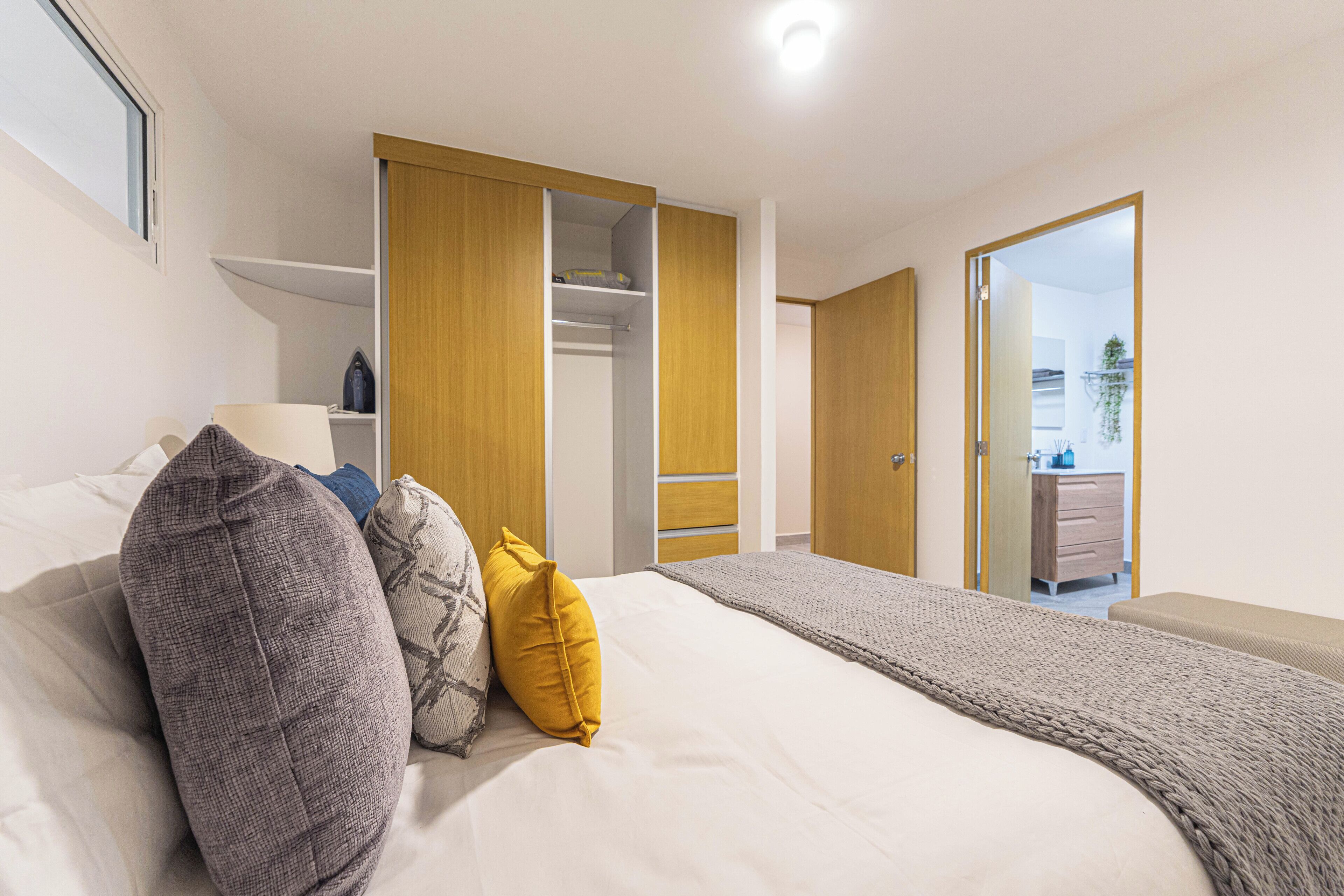 A bedroom with a bed, pillows, a bedside table, a wardrobe, and an open door leading to a bathroom.