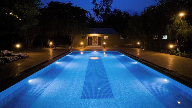 A swimming pool with a tiled deck, surrounded by trees and a building with a lighted entrance. - Tathastu Bandhavgarh (Manpur)
