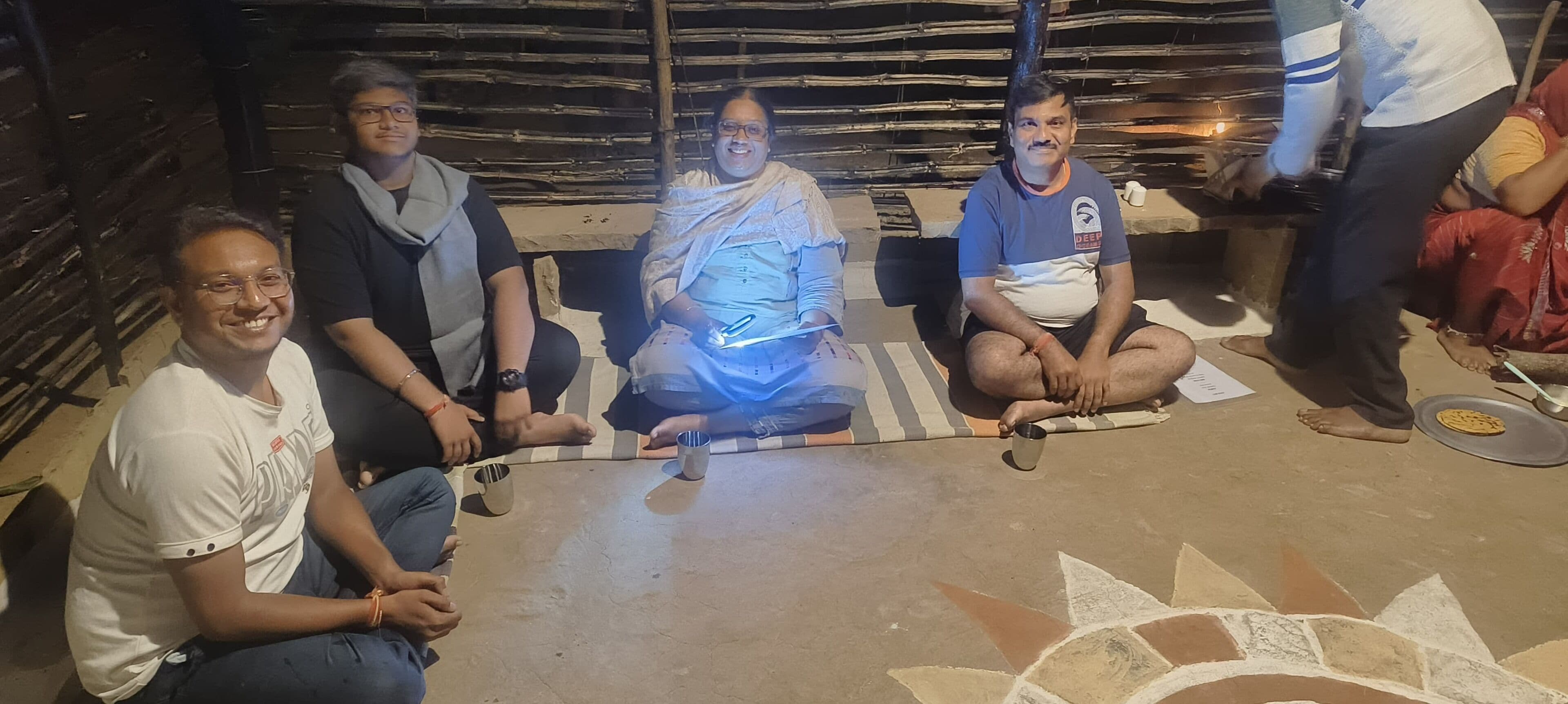 Un grupo de personas sentadas en un tapete en un entorno rústico con techo de paja y paredes de madera.