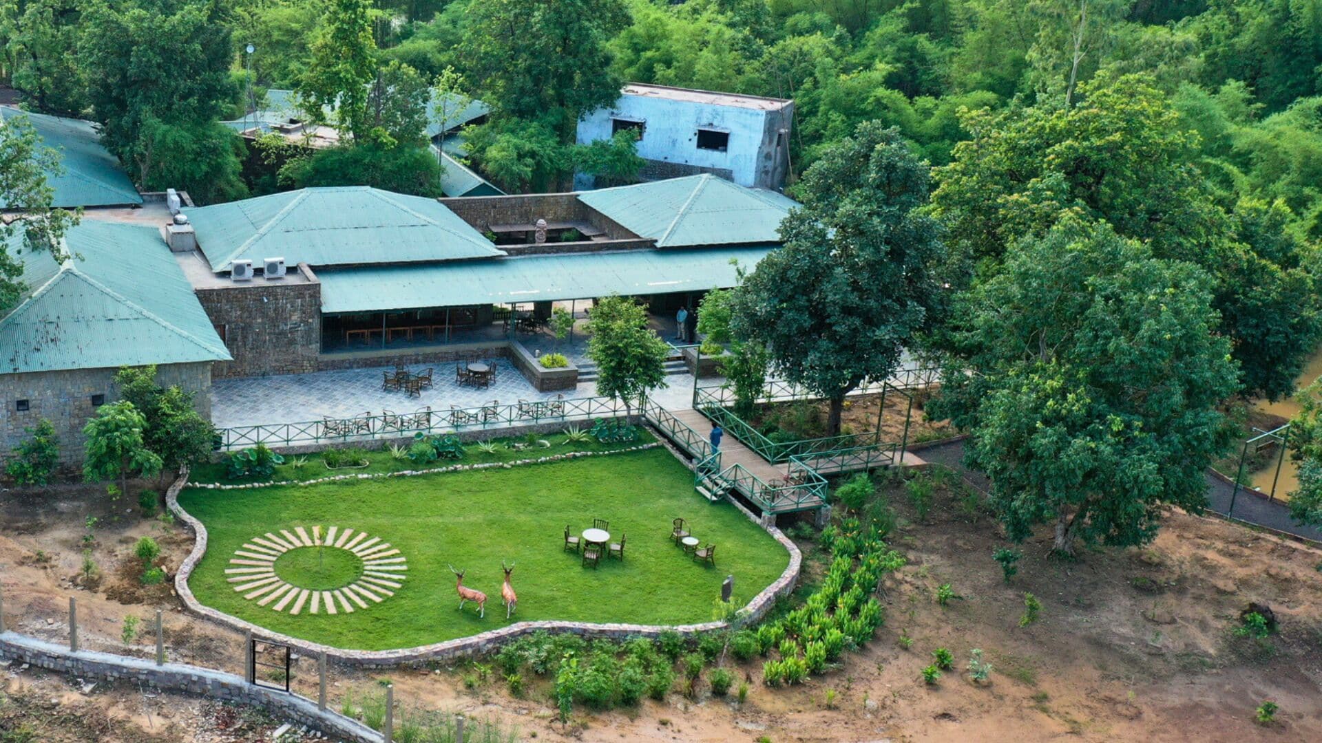 An aerial view of a building with a green roof, surrounded by lush greenery and a circular lawn with seating and two deer statues.