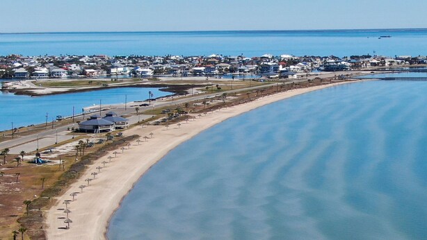 Vista aérea de una zona costera con una amplia playa de arena, aguas azules cristalinas y una zona residencial al fondo.