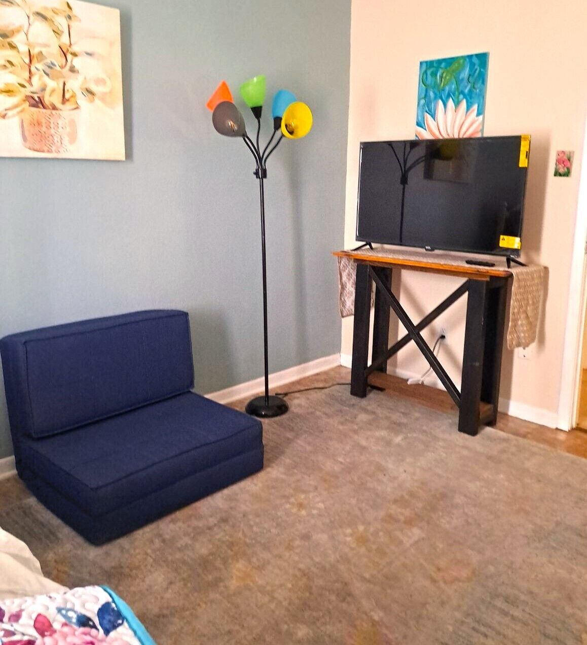 Una habitación con un sofá azul, una lámpara de pie, un televisor sobre un mueble de madera y un cuadro en la pared.