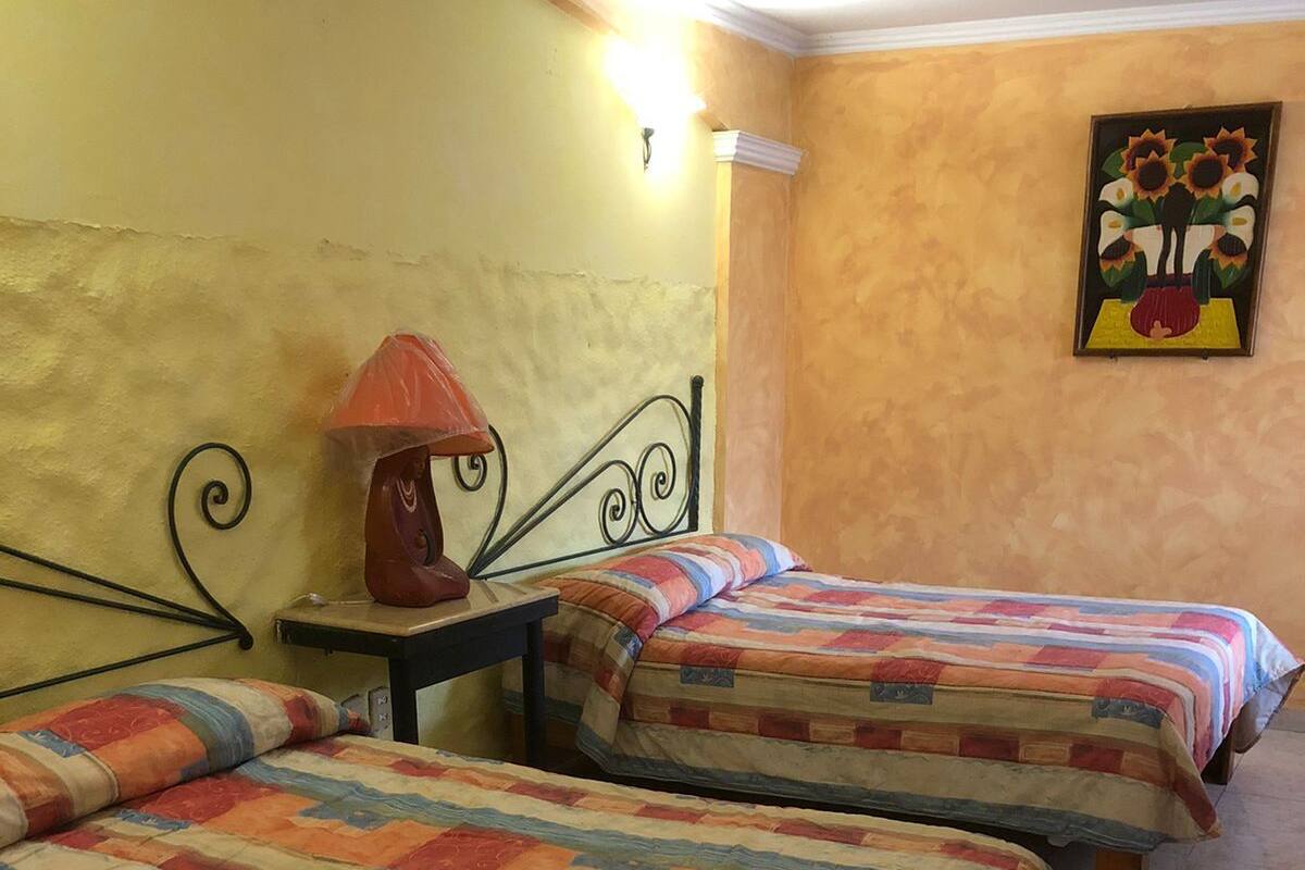 Ein Zimmer mit zwei Betten, jedes mit einer farbenfrohen Steudecke, einem kleinen Tisch mit einer Lampe und einem Bild an der Wand.